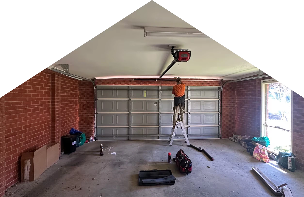 A person on a ladder repairs a gray garage door inside a garage with a red brick wall.